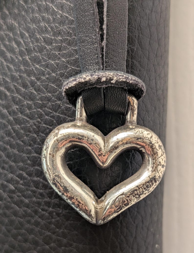May include: Close-up of a silver heart-shaped charm with the word "Brighton" engraved on it. The charm is attached to a black leather strap, set against a textured black leather background. The charm has a distressed, vintage appearance.