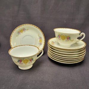 May include: A collection of vintage teacups and saucers. The white porcelain cups and saucers feature a floral design with yellow, pink, and blue flowers. Each piece has a gold trim around the edges. The set includes multiple cups and saucers.