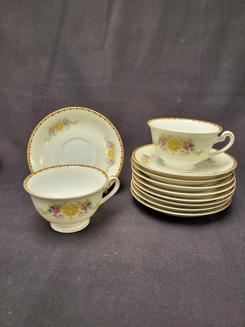 May include: A collection of vintage teacups and saucers. The white porcelain cups and saucers feature a floral design with yellow, pink, and blue flowers. Each piece has a gold trim around the edges. The set includes multiple cups and saucers.