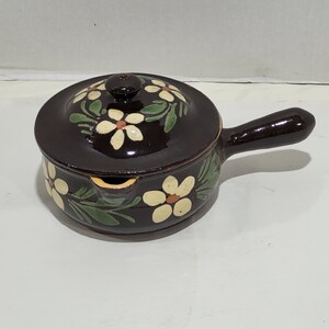 May include: A brown ceramic pot with a lid, featuring a white floral design with green leaves. The pot has a handle and a spout.