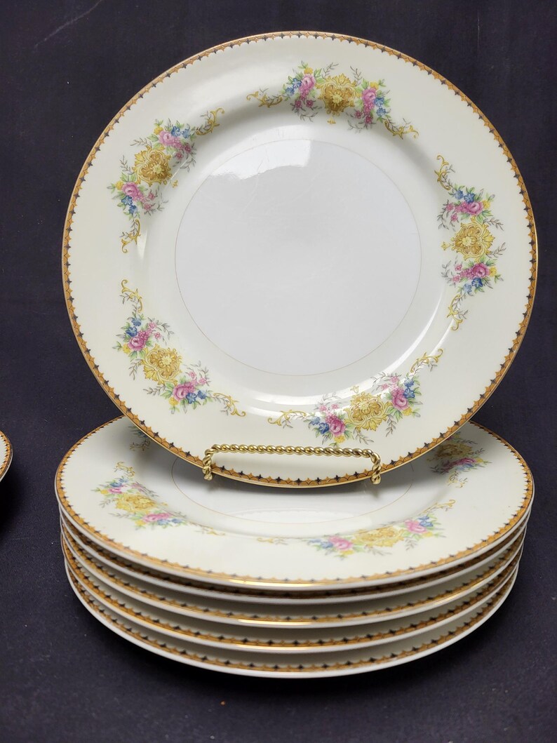 May include: A stack of white porcelain plates with a gold rim and floral design. The plates feature a gold border with a scalloped edge and a pattern of yellow, pink, and blue flowers. The plates are stacked on a dark surface.