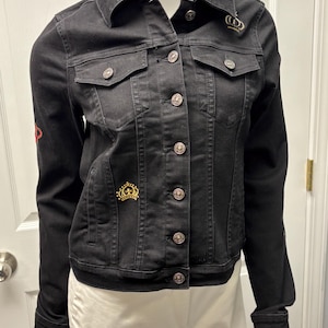 May include: Black denim jacket with silver buttons, two chest pockets, and a classic collar. Embellished with gold-colored crown pins on the chest and shoulder. The jacket has a tailored fit and long sleeves.