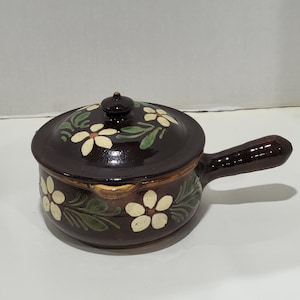 May include: A brown ceramic pot with a lid and a handle. The pot is decorated with white flowers and green leaves.
