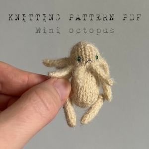 May include: A knitted mini octopus with green eyes. The octopus is beige and has a textured surface. The octopus is being held by a hand.