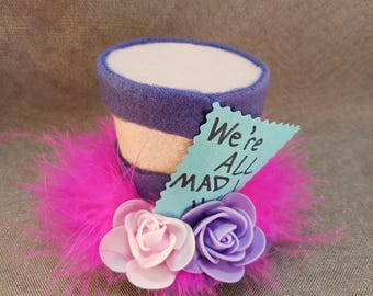 Pode incluir: Um chapéu alto de feltro roxo e branco com uma guarnição de penas rosa e duas rosas de tecido. O chapéu tem uma etiqueta azul anexada que diz "We're All Mad Here".