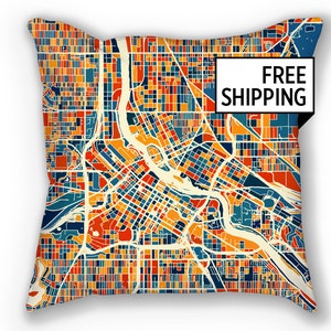 Può includere: Un cuscino decorativo colorato con una mappa stilizzata di una città. La mappa è in tonalità di rosso, arancione, giallo, blu e verde. Il cuscino è rettangolare e ha uno sfondo bianco. Il testo "FREE SHIPPING" è stampato su un rettangolo bianco nell'angolo in alto a destra del cuscino.