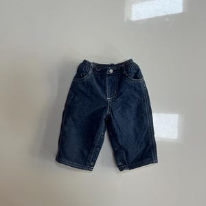 May include: Dark blue denim baby pants with an elastic waistband and snap closures on the sides. The pants have two front pockets and white stitching details. The garment is designed for infants or toddlers.