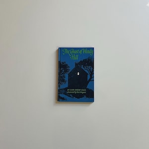 May include: A hardcover book titled "The Ghost of Windy Hill" with a blue cover. The cover features a silhouette of a house and trees against a dark blue background. The title is in green and white font.
