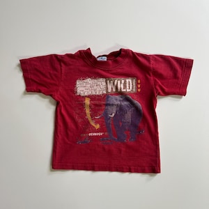 May include: Red t-shirt featuring a graphic of an elephant with the word "WILD" above it. The shirt also has the brand name "OSHKOSH" printed on it. The shirt is short-sleeved and has a crew neck.