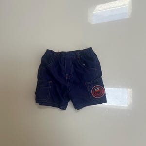 May include: Dark blue cargo shorts featuring a red Elmo character patch on the lower left pocket. The shorts have an elastic waistband, two side pockets, and visible red stitching details along the seams and pocket edges for a structured look.
