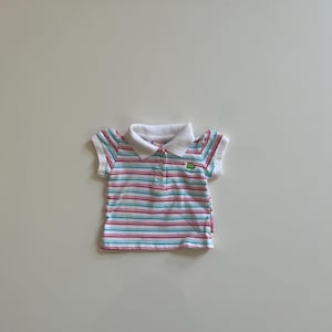 May include: A short-sleeved polo shirt with horizontal stripes in pink, blue, and white. The shirt has a white collar and sleeve cuffs. A small green embroidered design is on the chest.