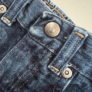 Op de afbeelding: Close-up van een blauwe denim jeans met zichtbare stiksels en metalen knoopsluitingen. De zilveren knopen zijn voorzien van de merknaam "Wrangler". De stof heeft een getextureerde uitstraling.