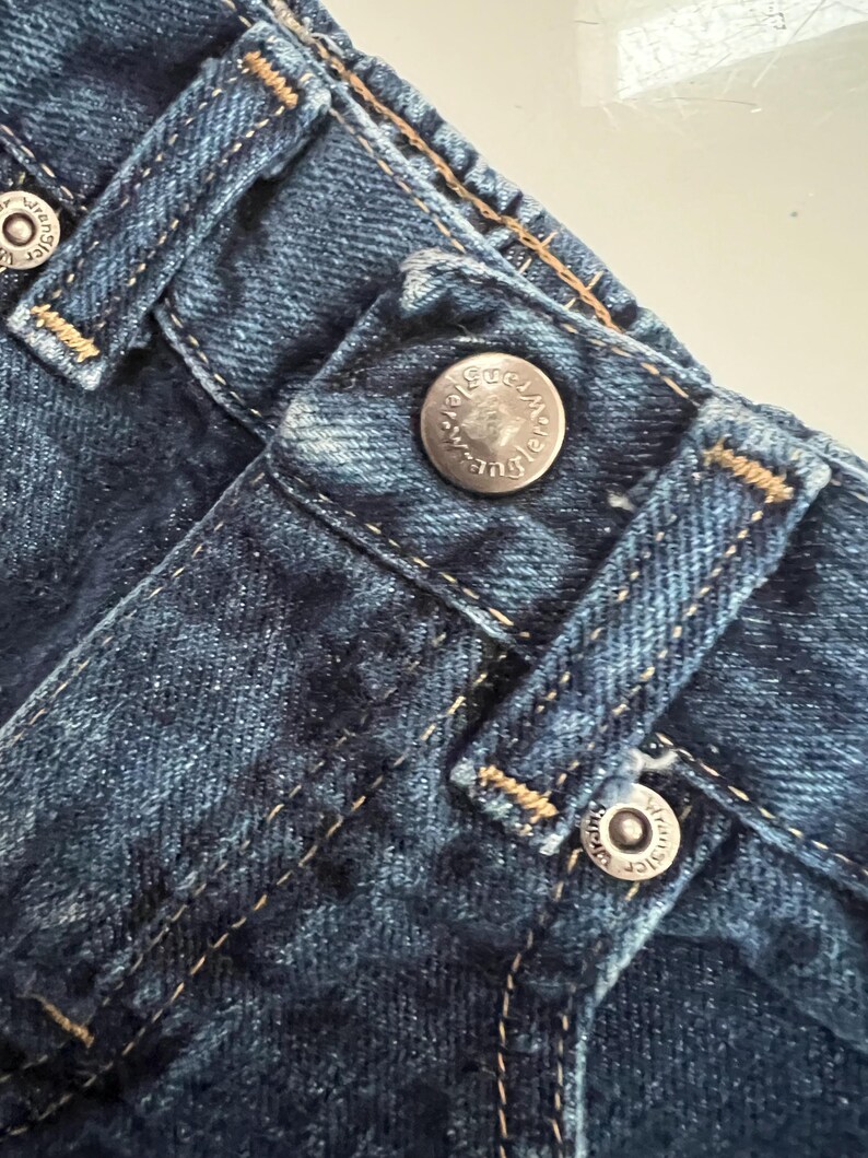 Op de afbeelding: Close-up van een blauwe denim jeans met zichtbare stiksels en metalen knoopsluitingen. De zilveren knopen zijn voorzien van de merknaam "Wrangler". De stof heeft een getextureerde uitstraling.