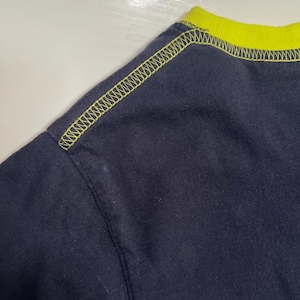 May include: A close-up of a navy blue t-shirt, featuring a neon yellow collar and stitching. The stitching is visible along the shoulder seam and collar, creating a contrasting design. The fabric appears to be a soft knit.