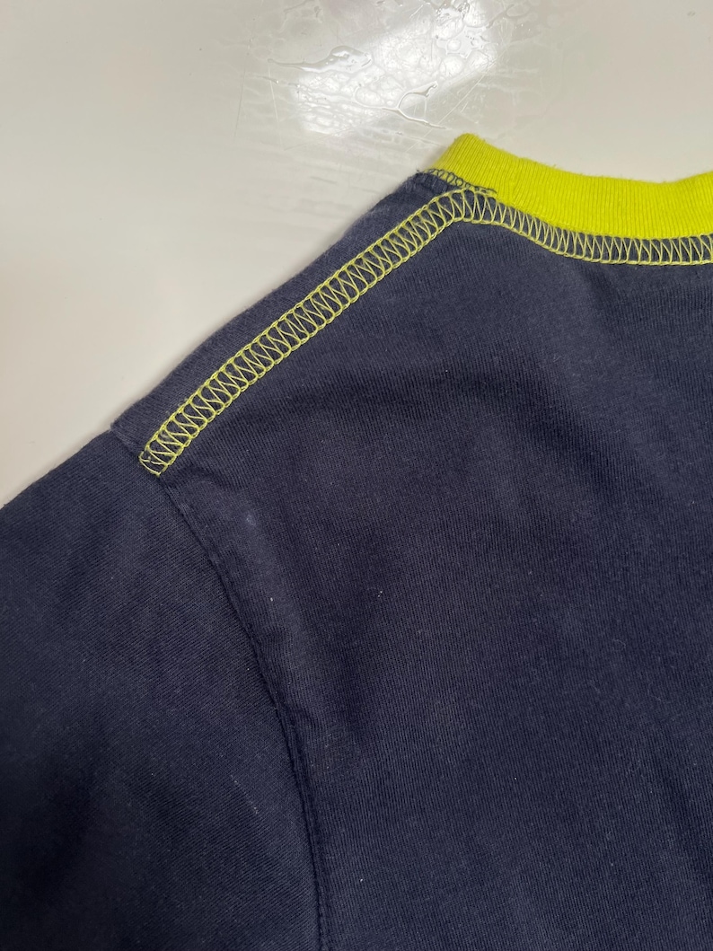 May include: A close-up of a navy blue t-shirt, featuring a neon yellow collar and stitching. The stitching is visible along the shoulder seam and collar, creating a contrasting design. The fabric appears to be a soft knit.