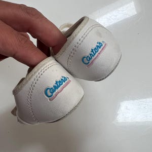 May include: A pair of white baby shoes with the brand name "Carter's" in blue script with a pink outline. The shoes have a rounded toe and a small lace. The shoes are made of a soft material.