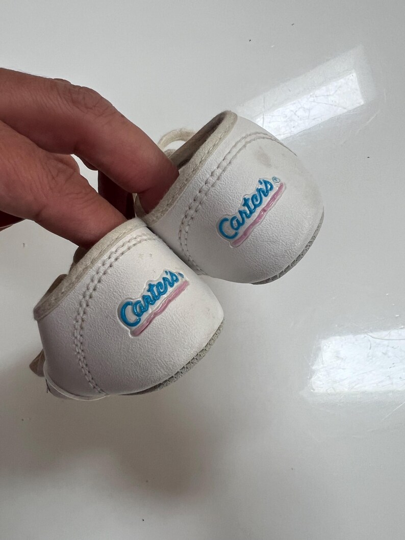 May include: A pair of white baby shoes with the brand name "Carter's" in blue script with a pink outline. The shoes have a rounded toe and a small lace. The shoes are made of a soft material.