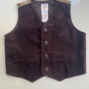 May include: A brown corduroy vest with a V-neck and three buttons. The vest has two front pockets and tan accents on the shoulders. The tag reads "GAP" and "5/m 8-15 lbs".