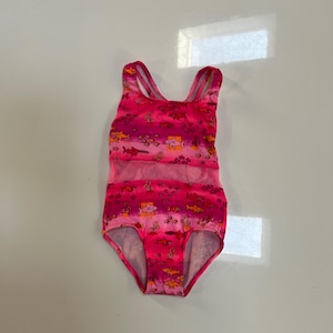 May include: A one-piece swimsuit in shades of bright pink and magenta, featuring a repeating pattern of orange and yellow sharks and fish. The garment has a scoop neckline, thick shoulder straps, and a horizontal mesh panel across the midsection.