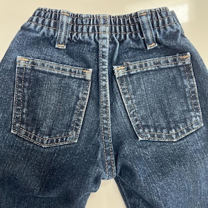 Op de afbeelding: Close-up van de achterkant van een donkerblauwe denim jeans. De jeans heeft twee achterzakken, een tailleband met riemlussen en een centrale gestikte naad. De stiksels zijn in een contrasterende bruine kleur.