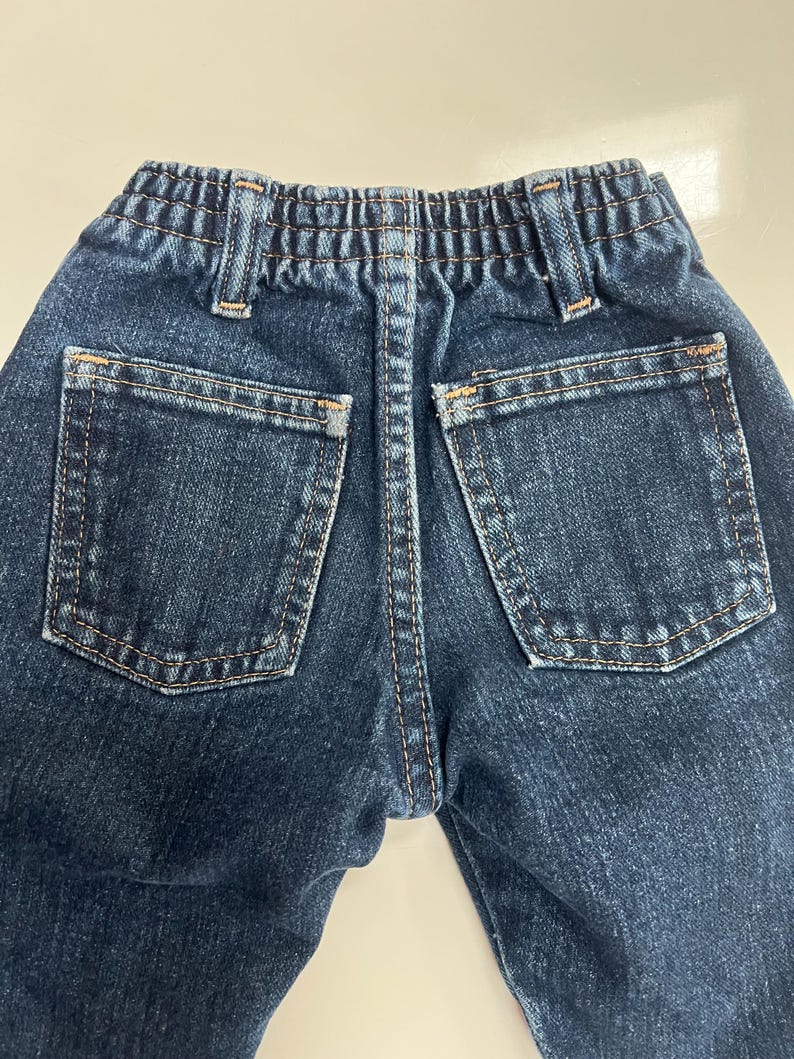 Op de afbeelding: Close-up van de achterkant van een donkerblauwe denim jeans. De jeans heeft twee achterzakken, een tailleband met riemlussen en een centrale gestikte naad. De stiksels zijn in een contrasterende bruine kleur.