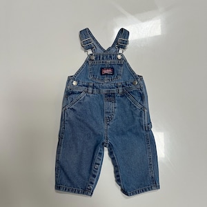 May include: Medium-wash blue denim overalls featuring adjustable metal buckle straps, a front bib pocket with a red and white OshKosh B'gosh logo patch, side button closures, and metal snap fasteners along the inner leg seams for easy access.