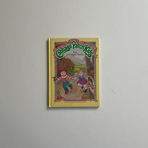 May include: A hardcover book titled "Cabbage Patch Kids: The Just-Right Family" with illustrations of children. The cover is yellow with green and red text and a gold border. A Parker Brothers story book.