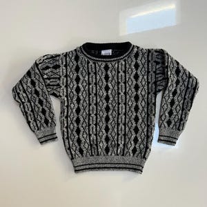 May include: A gray and black patterned sweater with a ribbed collar, cuffs, and hem. The sweater features a repeating diamond and vertical stripe design. The brand name "GARAY" is visible on the collar. The sweater is made of a soft, knitted material.
