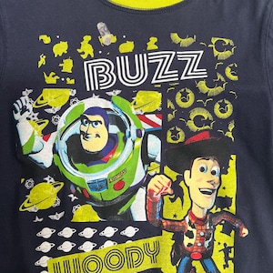 May include: Navy blue t-shirt with a yellow collar, featuring Buzz Lightyear and Woody from Toy Story. The shirt has the names "BUZZ" and "WOODY" in white and yellow. The design includes planets and other space-themed graphics.