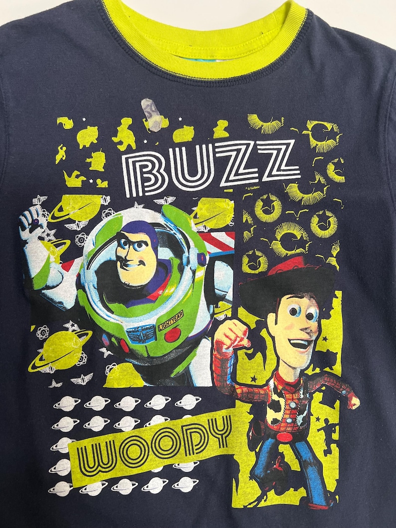 May include: Navy blue t-shirt with a yellow collar, featuring Buzz Lightyear and Woody from Toy Story. The shirt has the names "BUZZ" and "WOODY" in white and yellow. The design includes planets and other space-themed graphics.