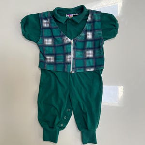 May include: A green baby romper with a collared shirt and plaid vest. The vest has a white zipper and a plaid pattern in shades of green, blue, and white. The romper has a button closure and elastic cuffs.