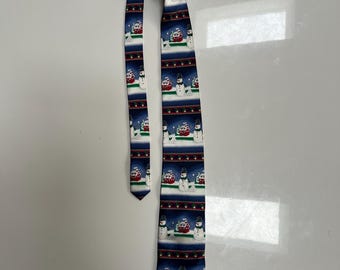 May include: A blue and white Christmas tie featuring a repeating pattern of snowmen, red houses, and snowflakes. The tie has a pointed end and is made of a smooth fabric. The background is a dark blue color.
