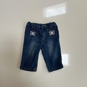May include: Dark wash denim baby jeans with embroidered butterfly details on the front pockets. The jeans have a button and zipper closure and are designed for comfort and style.