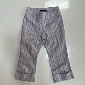May include: Capri pants featuring a vertical striped pattern in shades of light purple, white, and grey. The pants have a wide waistband with two light purple buttons, two front pockets, and cuffed hems at the bottom of each leg.