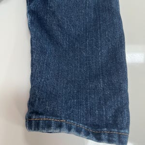 Op de afbeelding: Close-up van een deel van een blauwe spijkerbroek. De stof is een medium wassing met zichtbare textuur en subtiele kleurvariaties. De zoom is omgeslagen en vastgezet met contrasterende bruine stiksels.
