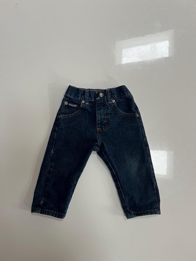 Op de afbeelding: Donkerblauwe spijkerbroek voor een peuter. De jeans heeft een knoop- en ritssluiting, voor- en achterzakken en het Wrangler-logo op de voorzak. De jeans is gemaakt van duurzaam denim materiaal.