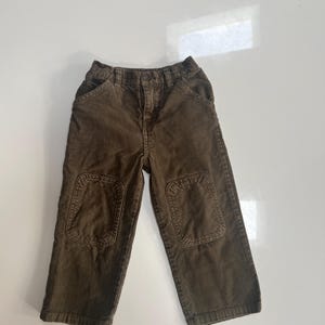 May include: Brown corduroy pants with reinforced knee patches. The pants have a classic five-pocket design and a button and zipper closure. The corduroy fabric has a ribbed texture. These pants are suitable for children.