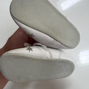 May include: A pair of white baby shoes with a star design. The shoes have a soft, gray sole and a small strap across the top. The shoes are made of a soft material.