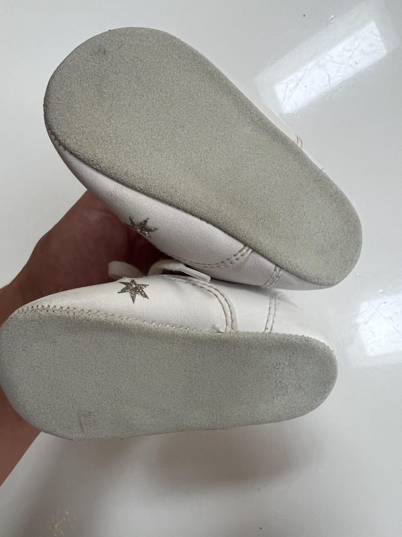 May include: A pair of white baby shoes with a star design. The shoes have a soft, gray sole and a small strap across the top. The shoes are made of a soft material.