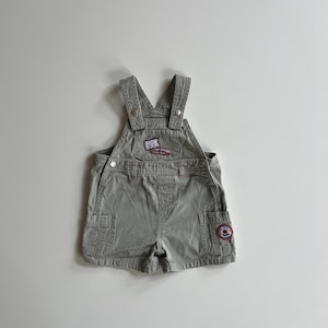 May include: Green overall shorts with a patch on the front and a red and white patch on the right leg.