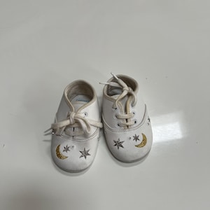 May include: A pair of white baby shoes with laces. Each shoe features a gold crescent moon and star design. The shoes are from the brand Carter's.