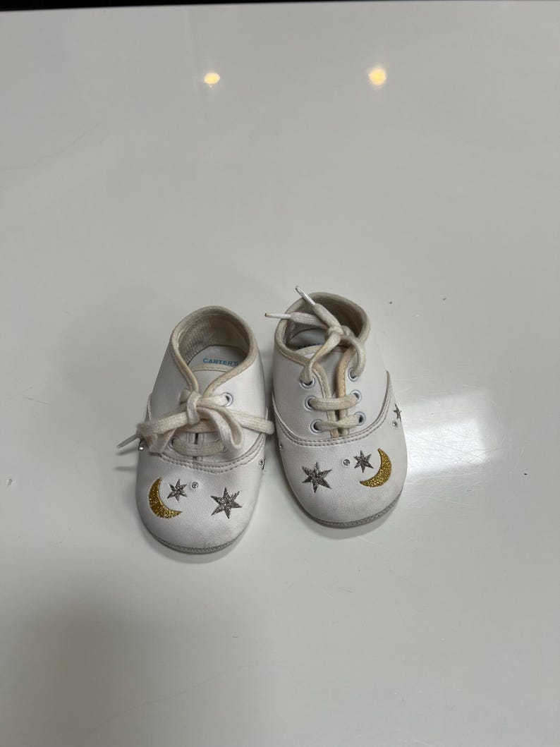 May include: A pair of white baby shoes with laces. Each shoe features a gold crescent moon and star design. The shoes are from the brand Carter's.