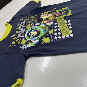 May include: Navy blue long-sleeved shirt with a yellow collar and cuffs. The shirt features a graphic of Buzz Lightyear and Woody from Toy Story, with the words "Buzz" and "Woody" in yellow.