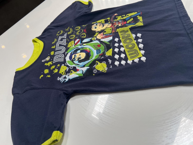 May include: Navy blue long-sleeved shirt with a yellow collar and cuffs. The shirt features a graphic of Buzz Lightyear and Woody from Toy Story, with the words "Buzz" and "Woody" in yellow.
