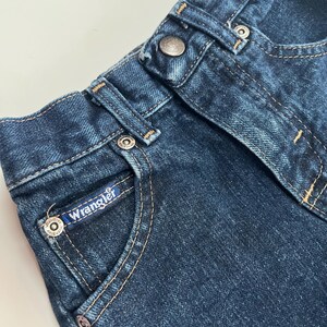Op de afbeelding: Close-up van een donkerblauwe denim jeans. De jeans heeft een knoopsluiting, riemlussen en een klein zakje met het "Wrangler"-logo. De stiksels zijn lichtbruin.