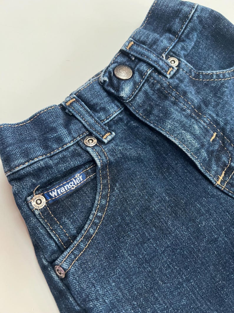 Op de afbeelding: Close-up van een donkerblauwe denim jeans. De jeans heeft een knoopsluiting, riemlussen en een klein zakje met het "Wrangler"-logo. De stiksels zijn lichtbruin.