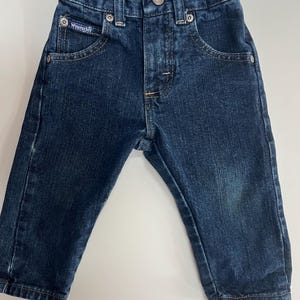 Op de afbeelding: Donkerblauwe Wrangler-jeans voor een baby of peuter. De jeans heeft een knoop- en ritssluiting, voor- en achterzakken en het Wrangler-logo op de voorzak.