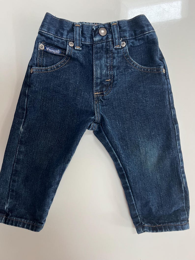 Op de afbeelding: Donkerblauwe Wrangler-jeans voor een baby of peuter. De jeans heeft een knoop- en ritssluiting, voor- en achterzakken en het Wrangler-logo op de voorzak.