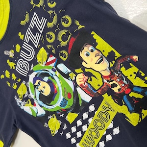 May include: Navy blue t-shirt with a yellow collar and trim. The shirt features a graphic of Buzz Lightyear in his spaceship and Woody from Toy Story. The word "BUZZ" is printed above Buzz, and "WOODY" is printed below Woody.