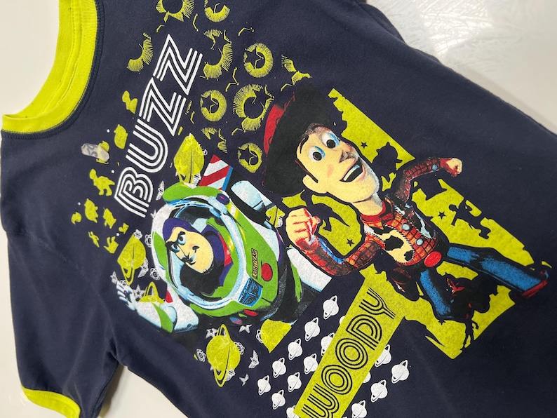 May include: Navy blue t-shirt with a yellow collar and trim. The shirt features a graphic of Buzz Lightyear in his spaceship and Woody from Toy Story. The word "BUZZ" is printed above Buzz, and "WOODY" is printed below Woody.
